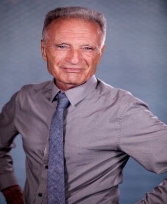 A confident man in a grey shirt and tie, smiling and standing with his hands on his hips against a neutral background.