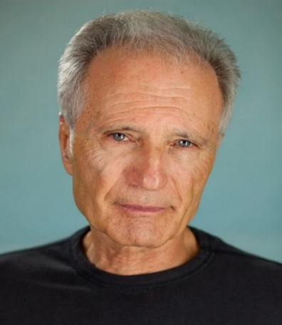 An older man with short gray hair, wearing a black shirt, looking serious against a neutral background.